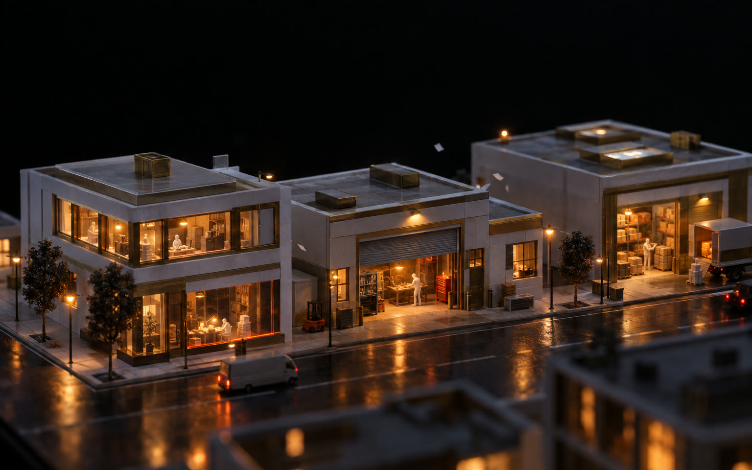 A detailed model of a small main street at night — an office, a workshop and a warehouse — every window lit warm.